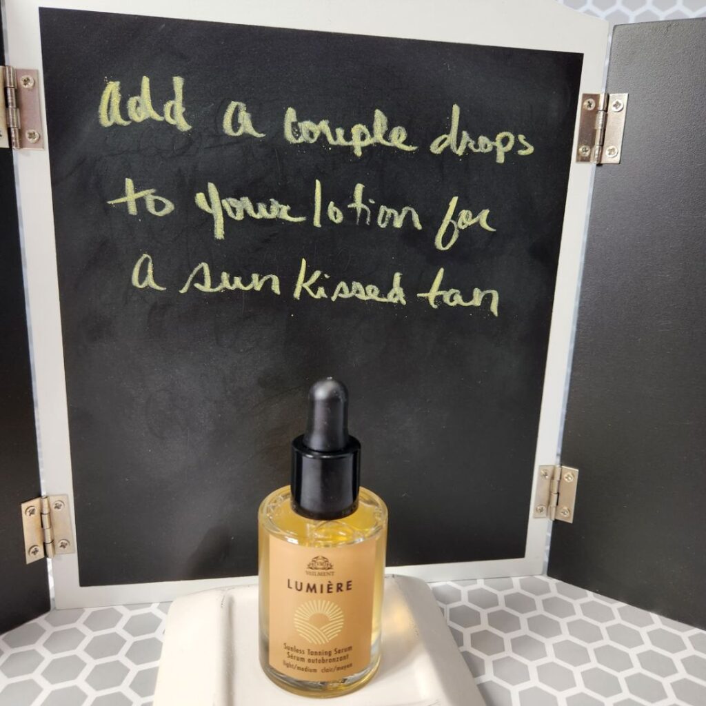 bottle of Lumiere Sunless Tanning Serum in front of blackboard that reads: Add a couple drops to your lotion for a sun kissed tan
