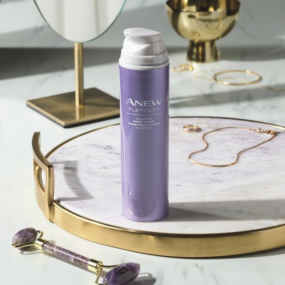 bottle of Anew Platinum Sculpting Body Serum on a marble and gold tray on a marble counter with a few pieces of simple gold jewelry, a stand mirror and purple stone roller tool